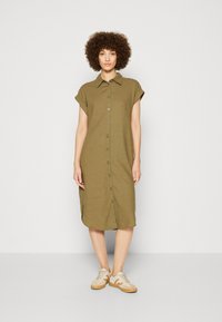 Olivgrön linneskjortklänning med korta uppvikta ärmar, knappstängning fram, rundad nederkant och fickor. Matchas med beige sneakers.