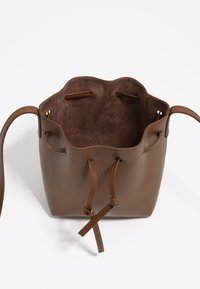 Hnedá kožená taška typu bucket s hladkým exteriérom, vlnitým horným okrajom a interiérom s zamakou. Obsahuje sťahovaciu šnúrku a nastaviteľné popruhy.