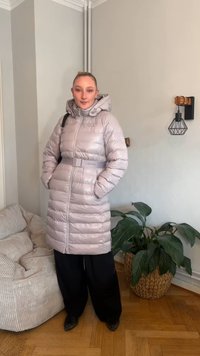 Dolga, svetlo roza podložena jakna s kapuco, pasom in sijočim zaključkom. Nošena preko črnih hlač širokih nog, stoječa v udobnem notranjem prostoru.