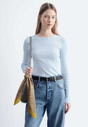 Jeune femme portant un haut bleu clair à manches longues et un jean bleu tient un sac à main gris avec une écharpe jaune à motifs attachée à la bandoulière.