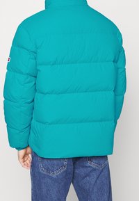 Teal puffjacka med horisontella quiltade sektioner, hög krage och elastiska ärmslut. Har en logotyp på vänster ärm.
