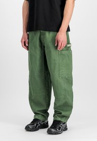 Zöld cargo nadrág, puha anyagból készült, több zsebbel, részletes varrásokkal és laza szabással, fekete sportcipőkkel párosítva.