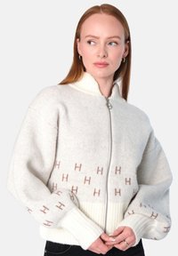 Ullzippsweater i lys beige med ribbet hvit nederkant og mansjetter. Har et subtilt logomønster i lys brun på kroppen.