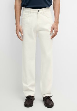 Mann trägt gerade geschnittene weiße Hose, dunkelbraune Leder-Schnürschuhe und ein dunkelblaues Hemd, steht vor einem einfarbigen Hintergrund.