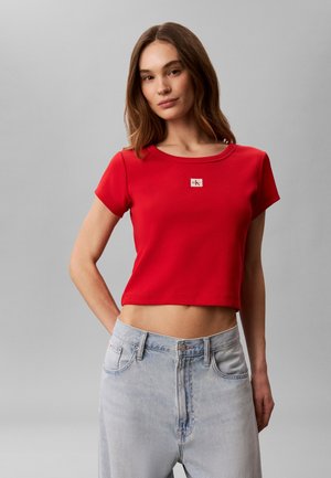 Νεαρή γυναίκα με καστανά μαλλιά που φοράει κόκκινη κοντή μπλούζα με μικρό λογότυπο και ανοιχτό μπλε ψηλόμεσο τζιν με γκρι φόντο.