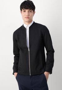 Μαύρο μπουφάν με φερμουάρ κατασκευασμένο από μαλακό ύφασμα, με στρογγυλή λαιμόκοψη, μακρυά μανίκια και λεία υφή. Φοριέται πάνω από ένα άσπρο πουκάμισο.