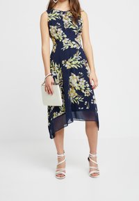 Bloemen navy jurk met een strakke lijfje en een vloeiende rok, met gele en witte bloemenpatronen. Gecombineerd met een kleine witte handtas en sandalen met banden.