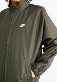Zelena vetrovka olivno zelene barve z visokim ovratnikom, elastičnimi manšetami in stranskimi žepi. Na levi strani prsnega dela je bel Nike logo.