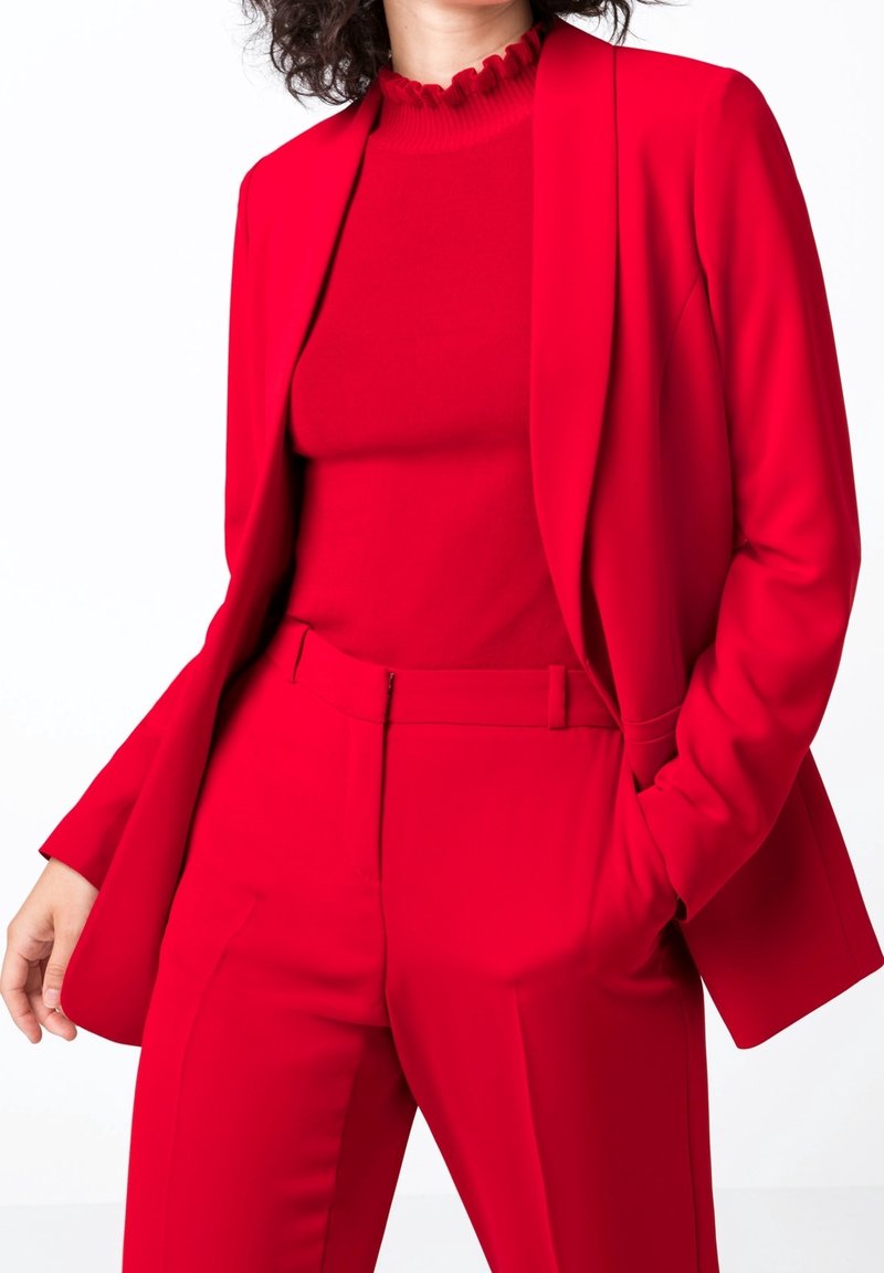Femme portant un tailleur rouge vif sur mesure avec un blazer, un pantalon taille haute et un haut à col volanté, debout avec une main dans la poche.