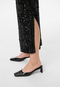 Crna haljina s šljokicama i bočnim prorezom, uparen sa crnim lakiranim mule sandalama sa kvadratnim vrhom i niskom, debelom potpeticom. Glatka tekstura i sjajni završetak.