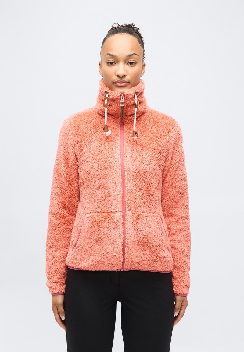 Γυναίκα που φοράει φλις σακάκι κοραλλί ροζ με ψηλό γιακά με φερμουάρ και κορδόνια, στέκεται μπροστά σε ένα απλό φωτεινό φόντο.