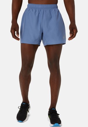 Mann trägt hellblaue Sportshorts und schwarze Laufschuhe mit blauen Akzenten, steht vor einem schlichten hellen Hintergrund.