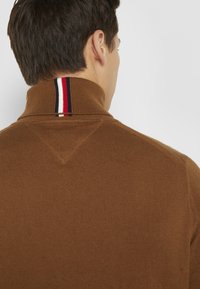 Brun ribstrikket sweater med høj krave og en rød, hvid og blå stribe detalje bagpå. Tekstureret stof med en struktureret pasform.