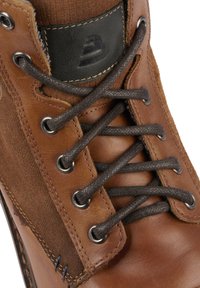 Hnědé kožené boty s tmavě hnědými semišovými akcenty, kulatými kovovými očky, černou logovou nášivkou a plochými tkaničkami v křížovém vzoru.