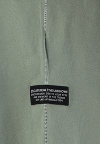 Tela verde con un acabado texturizado, que presenta una etiqueta negra con texto blanco y costuras de contraste visibles a lo largo del borde.