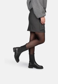 Botas de tobillo de cuero negro con un acabado texturizado, paneles elásticos laterales y un tacón bajo cuadrado, combinadas con una falda gris y medias negras.