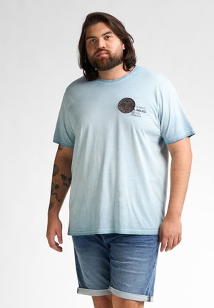 T-shirt en coton bleu clair avec un col rond, des manches courtes et un petit patch graphique sur la poitrine. Associé à un short en denim.