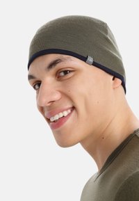 Olivově zelená pletená čepice s námořnickou modrou krempou a malou značka po straně. Přiléhavý design se přizpůsobuje tvaru hlavy.