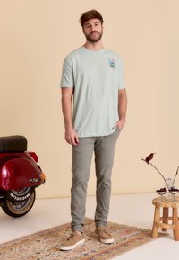 Lichtgroene t-shirt met korte mouwen met een kleurrafische palmboom. Gecombineerd met lichtgrijze broek en beige veterschoenen. De achtergrond bevat een vintage scooter.