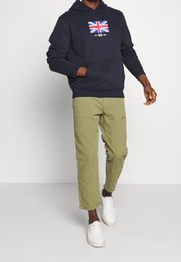 Man in een marineblauwe hoodie met VK-vlag grafiek, olijfgroene broek met patchzakken en witte sneakers, die tegen een effen achtergrond loopt.