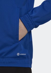 Blå idrottsjacka i texturerad tyg, med sidoficka och ribbade muddar. Har "AEROREADY"-logotyp vid fållen.