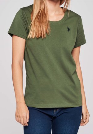 Vrouw draagt een groen T-shirt met korte mouwen en een ronde hals, met een klein zwart logo op de bovenborst, gecombineerd met donkerblauwe jeans.