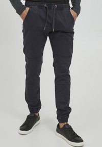 Navy sweatpants with elastic cuffs and drawstring waist, featuring side pockets and stitched panels for added detail, paired with black sneakers.