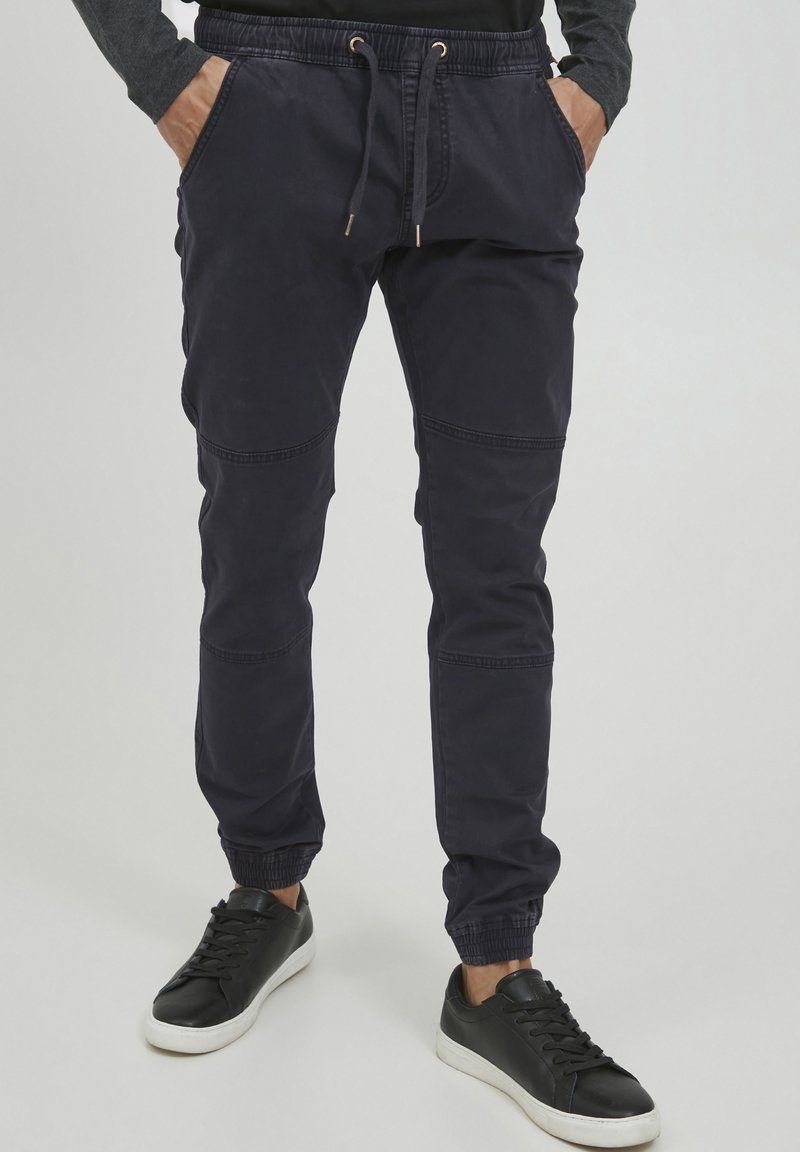 Marineblå joggebukser med elastiske mansjetter og snøring i livet, med side lommer og sydde paneler for ekstra detaljer, kombinert med svarte joggesko.