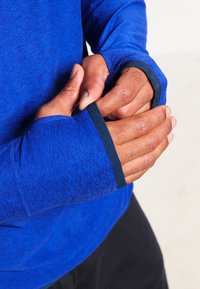 Långärmad träningsskjorta i heathered blått tyg, med tumhål och svart kant på ärmsluten. Slät textur och figurnära design.