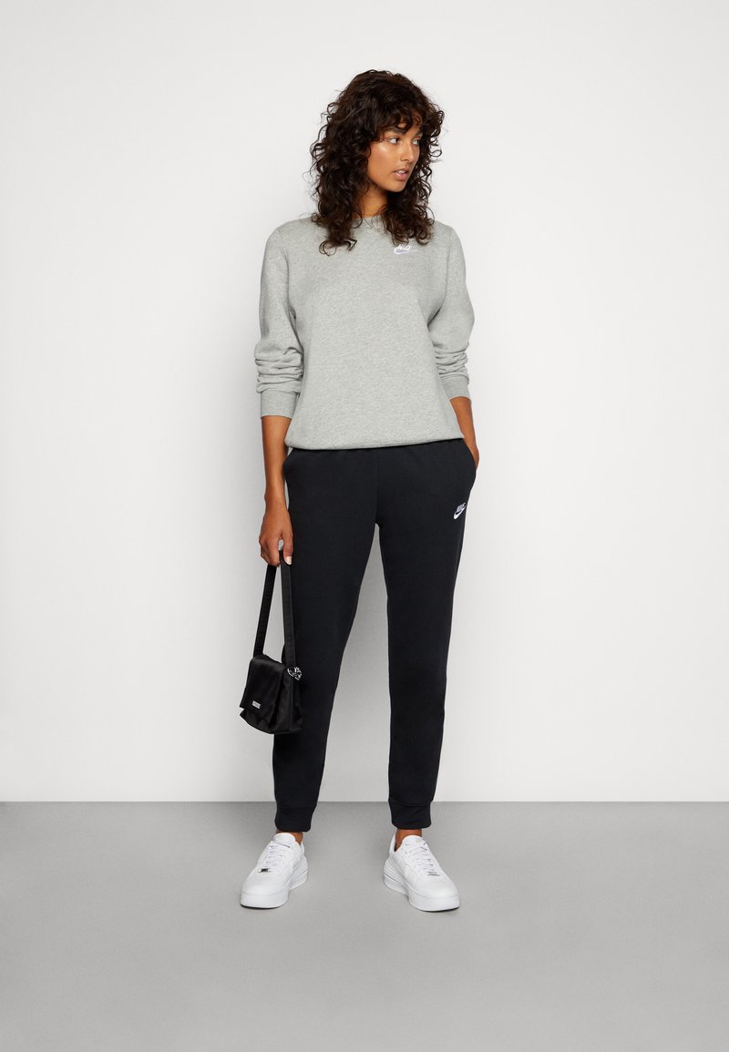 Sweatshirt gris avec des poignets côtelés, associé à un pantalon de jogging noir arborant un logo, complété par des baskets blanches et un petit sac noir.