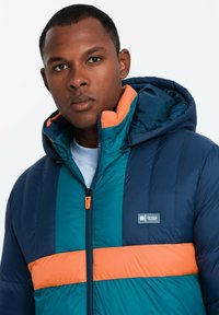 Vadderad jacka med huva, i marinblått, teal och orange paneler. Slät textur, synlig dragkedja och logotyp på bröstet.