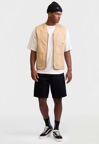 Beżowa kamizelka z płótna z dwoma przednimi kieszeniami, zapinana na zamek, noszona na białej koszulce, w połączeniu z czarnymi szortami i czarnymi trampkami.