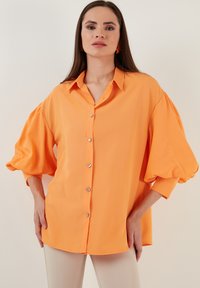 Oranžs krekls ar pogām un platām, pūkainām piedurknēm, brīva piegriezuma. Gluda auduma, spīdīgas pogas un atvieglināts apkakle.