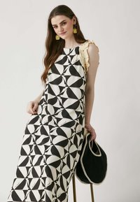 Maxi abito con motivo geometrico in bianco e nero, maniche leggere a volant, abbinato a una borsa a tracolla nera. Tessuto dalla trama morbida.