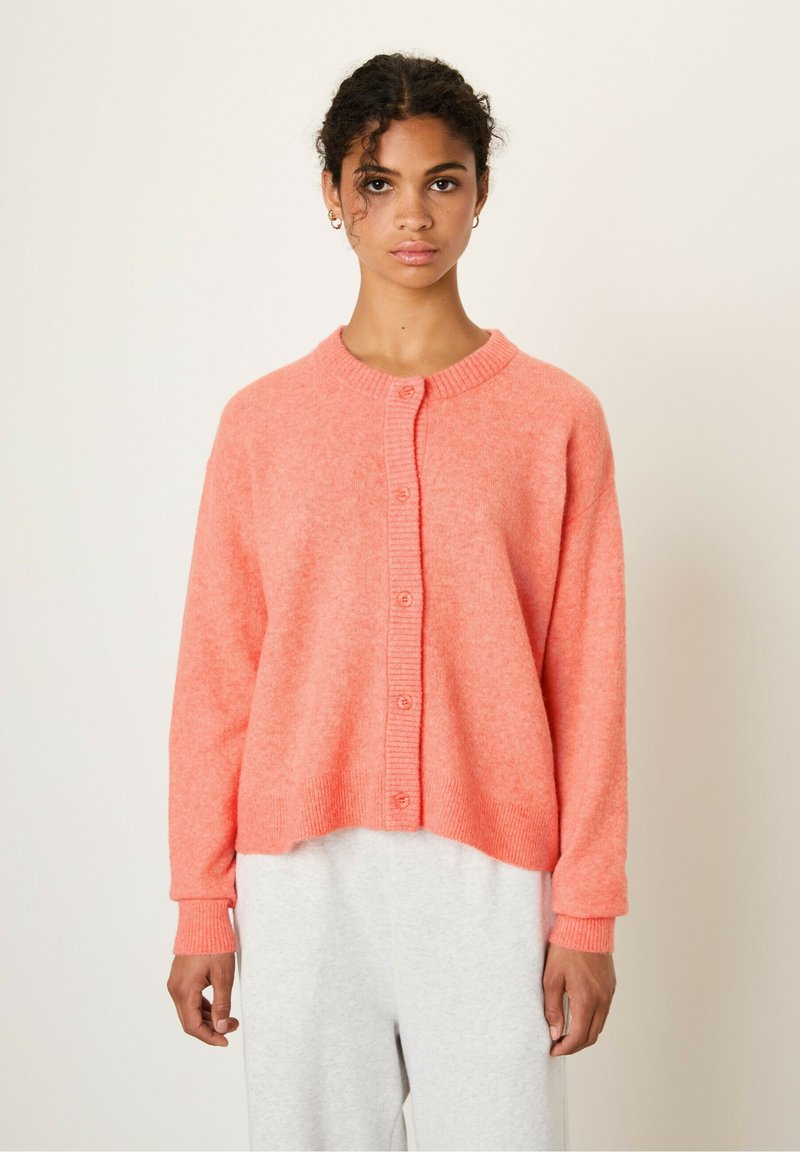 Koralni kardigan iz mehkega materiala. Ima okroglo izrezano ovratnico, zapenjanje na gumb in rebraste manšete, s sproščenim, skrajšanim krojem.