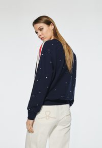 Marinblå sweatshirt med silverdetaljer, långa ärmar och röda samt creamfärgade ränder längs axlarna. Parat med vita byxor.