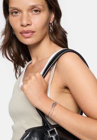 Silverfärgad kedjearmband med en knuten detalj på handleden. Vit och beige tanktop, håller en svart läderhandväska med texturerad yta.