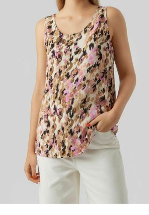 Mouwloze bloemen top in beige, roze en zwarte tinten, gedragen met witte broek, één hand in de zak, neutrale grijze achtergrond.