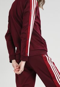 Vinröd idrottsbyxa med vit tre-strecksdesign längs ärmar och byxor. Tyget ser slätt ut med tryckknappspynt vid ärmsluten.