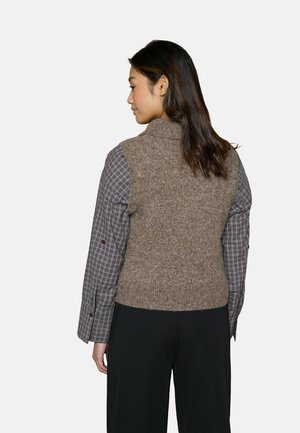 Brun vest i uldblanding med høj krave, båret over en ternet skjorte. Har en struktureret finish og tætsiddende design, kombineret med sorte bukser.