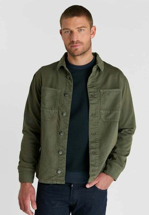 Man met kort haar die een open groene jas draagt over een donkere trui en donkere broek, staand voor een effen lichte achtergrond.