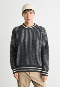 Strikkegenser i mørkegrått med et mønsterdesign, ribbet rund hals og kontrasterende hvite striper i kanten og ved ermer. Akkompagnert av en beige lue.