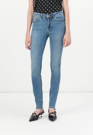 Frau trägt hellblaue Skinny-Jeans, schwarze Lack-Pumps mit spitzer Spitze und ein ärmelloses schwarzes Oberteil mit weißen Punkten vor weißem Hintergrund.