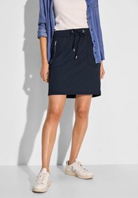 Un short-jupe bleu marine avec une ceinture à cordon de serrage, doté de deux poches zippées et d'une texture lisse, porté avec une chemise claire et des baskets blanches.