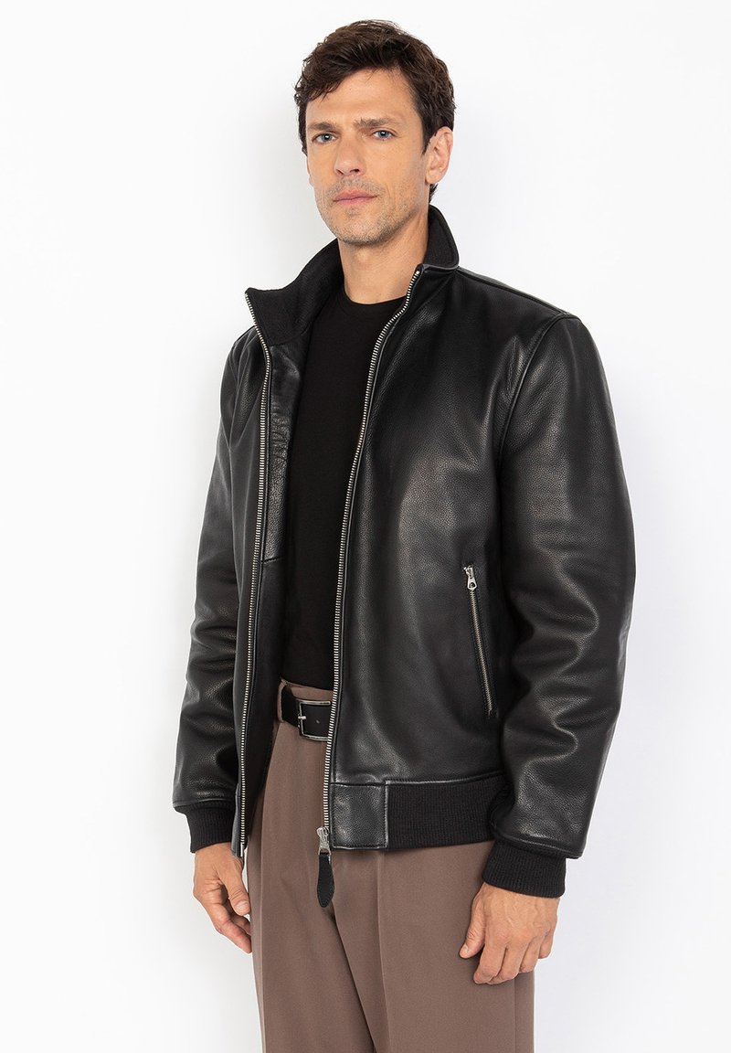 Schwarzleder-Bomberjacke mit Stehkragen, Frontreißverschluss, zwei Seiten pockets und gerippten Bündchen und Saum, getragen über einem schwarzen Hemd.