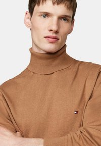 Brauner Rollkragenpullover mit gerippter Textur, versehen mit einem kleinen Logo-Patch auf der Brust. Schlichtes Design mit einer figurschmeichelnden Silhouette.