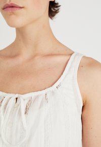 Witte mouwloze top van een zachte, gestructureerde stof. Voorzien van een kantafwerking, een delicate knoopsluiting en een trekkoord bij de hals.