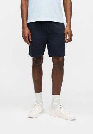 Homme portant un short bleu marine, des chaussettes blanches, des baskets blanches et une chemise bleu clair, debout devant un fond blanc uni.