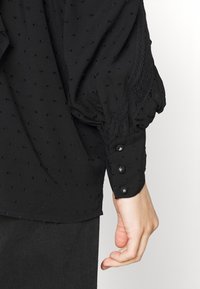 Blusa nera con pois texturizzati, maniche a sbuffo, dettagli in pizzo e chiusure decorative con bottoni lungo i polsini. Tessuto morbido e leggero.