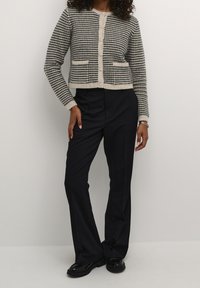 Cardigan a quadri grigio e beige con scollatura rotonda, chiusura a bottoni e due tasche frontali, abbinato a pantaloni neri su misura e scarpe.